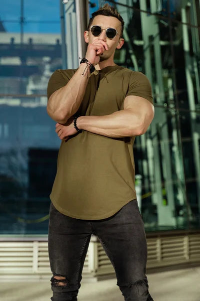 Ein Mann trägt ein armeegrünes T-Shirt, zerrissene schwarze Jeans und eine Sonnenbrille, mit einem Glasgebäude im Hintergrund.