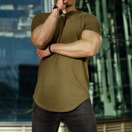 Ein Mann trägt ein armeegrünes T-Shirt, zerrissene schwarze Jeans und eine Sonnenbrille, mit einem Glasgebäude im Hintergrund.