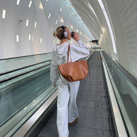 Eine frau in einem grauen outfit und kopfhörern geht durch einen modernen flughafenflur und trägt eine braun ledertasche