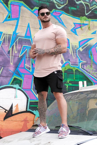 Ein Mann steht vor einer mit farbenfrohen Graffiti bedeckten Wand und trägt ein rosa T-Shirt, schwarze Shorts und Turnschuhe.