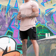 Ein Mann steht vor einer mit farbenfrohen Graffiti bedeckten Wand und trägt ein rosa T-Shirt, schwarze Shorts und Turnschuhe.