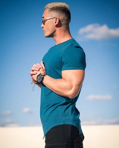 Seitenansicht eines Mannes, der ein blaugrünes T-Shirt trägt und im Freien mit einem blauen Himmel und Wolken im Hintergrund steht.