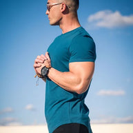 Seitenansicht eines Mannes, der ein blaugrünes T-Shirt trägt und im Freien mit einem blauen Himmel und Wolken im Hintergrund steht.