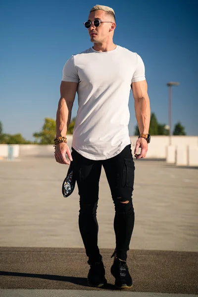 Ein Mann mit weißem T-Shirt, schwarzen Jeans und schwarzen Turnschuhen steht im Freien unter einem klaren blauen Himmel.