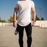 Ein Mann mit weißem T-Shirt, schwarzen Jeans und schwarzen Turnschuhen steht im Freien unter einem klaren blauen Himmel.