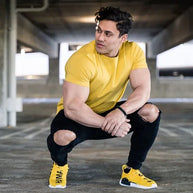 Mann trägt ein gelbes T-Shirt, dazu eine schwarze Hose und gelbe Turnschuhe, sitzt in geduckter Position