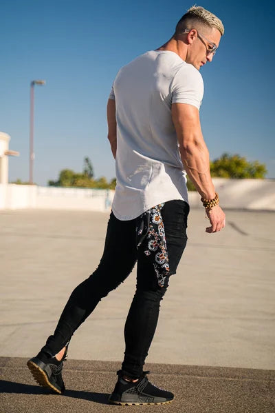 Ein Mann mit weißem T-Shirt, schwarzen Jeans und schwarzen Turnschuhen steht im Freien unter einem klaren blauen Himmel.