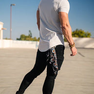 Ein Mann mit weißem T-Shirt, schwarzen Jeans und schwarzen Turnschuhen steht im Freien unter einem klaren blauen Himmel.