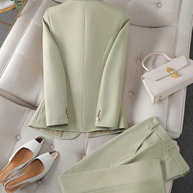 Ein stylischer hellgrüner Blazer und eine passende Hose auf neutralem Untergrund, dazu eine kleine Handtasche und ein Paar weiße Sandalen.