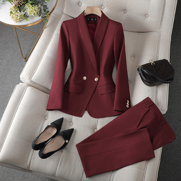 Ein burgunderroter Blazer und eine passende Hose auf einer weichen Oberfläche, kombiniert mit schwarzen Absätzen und einer kleinen schwarzen Handtasche.