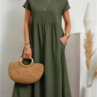Frau in einem dunkelgrünen Sommerkleid hält eine Tasche und steht vor einer weißen Wand.