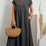 Stilvolle Frau in einem dunkelgrauen Sommerkleid mit einer Tasche vor einer weißen Wand.