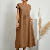 Frau in einem braunen, fließenden Sommerkleid steht vor einer weißen Wand