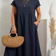 Frau in einem marineblauen Sommerkleid hält eine Tasche und steht vor einer weißen Wand.