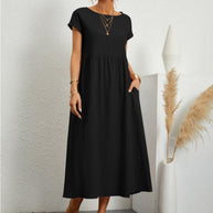 Frau in schwarzem Sommerkleid, eine Hand auf dem Po, steht vor weißer Wand