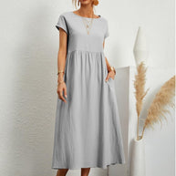 Frau in silbergrauem Sommerkleid, eine Hand in der Tasche, steht vor weißer Wand